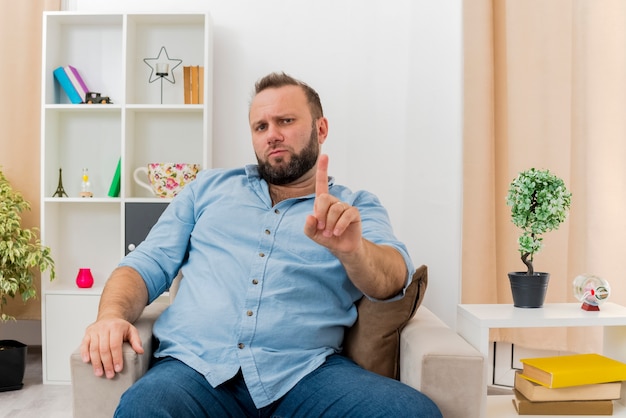 Hombre eslavo adulto confiado se sienta en un sillón mostrando el dedo índice mirando dentro de la sala de estar diseñada