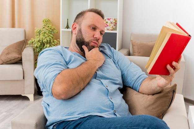 Hombre eslavo adulto confiado se sienta en el sillón poniendo la mano en la barbilla sosteniendo y mirando el libro dentro de la sala de estar