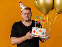 Foto gratuita hombre eslavo adulto decepcionado con gafas ópticas con gorro de cumpleaños sostiene globos de helio y pastel de cumpleaños