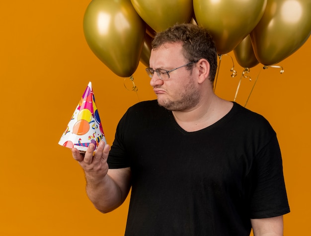 Foto gratuita hombre eslavo adulto decepcionado con gafas ópticas sosteniendo y mirando la gorra de cumpleaños se encuentra frente a globos de helio
