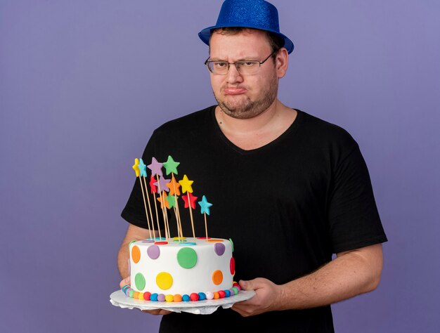 Hombre eslavo adulto disgustado con gafas ópticas vistiendo gorro de fiesta azul tiene pastel de cumpleaños
