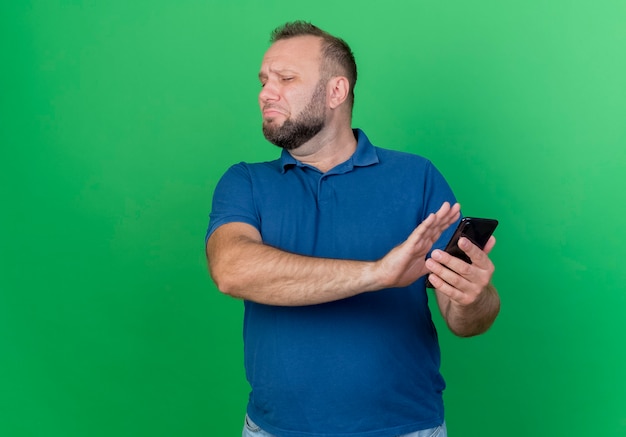 Hombre eslavo adulto disgustado que sostiene el teléfono móvil y no hace ningún gesto con la mano aislada en la pared verde con espacio de copia