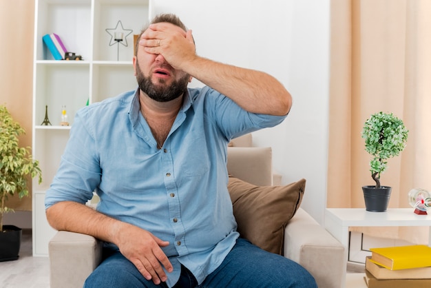 Hombre eslavo adulto disgustado se sienta en un sillón poniendo la mano en la cara cerrando los ojos dentro de la sala de estar