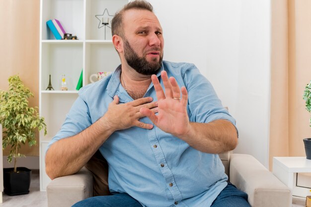 Hombre eslavo adulto disgustado se sienta en un sillón poniendo la mano en el pecho y no gesticula ningún signo de mano dentro de la sala de estar
