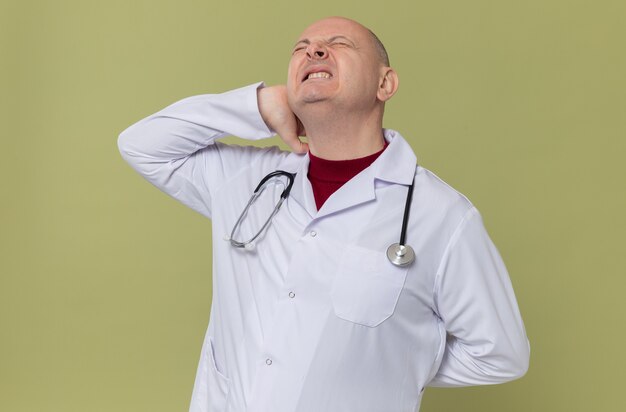 Hombre eslavo adulto dolorido en uniforme médico con estetoscopio poniendo la mano en el cuello y reteniendo