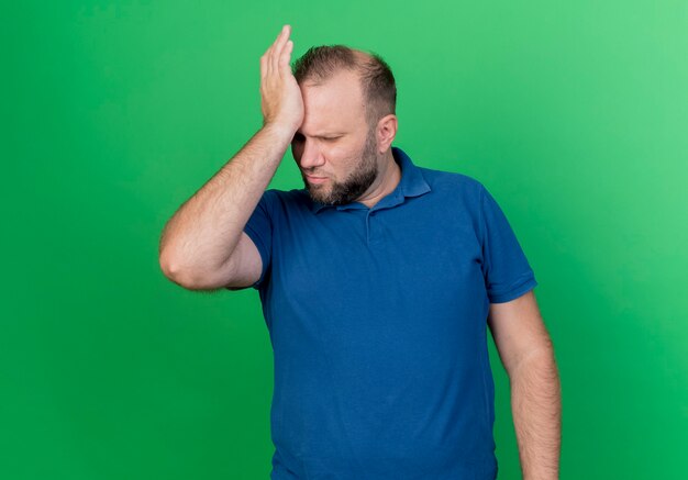 Hombre eslavo adulto frunciendo el ceño poniendo la mano en la cabeza mirando hacia abajo que sufre de dolor de cabeza aislado en la pared verde con espacio de copia