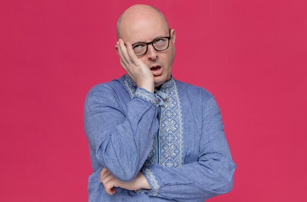 Hombre eslavo adulto molesto en camisa azul con gafas ópticas poniendo la mano en la cara y poniendo los ojos en blanco