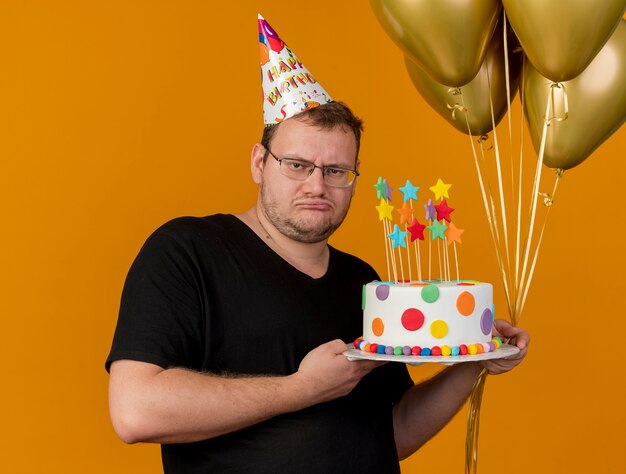 Hombre eslavo adulto molesto con gafas ópticas con gorro de cumpleaños sostiene globos de helio y pastel de cumpleaños