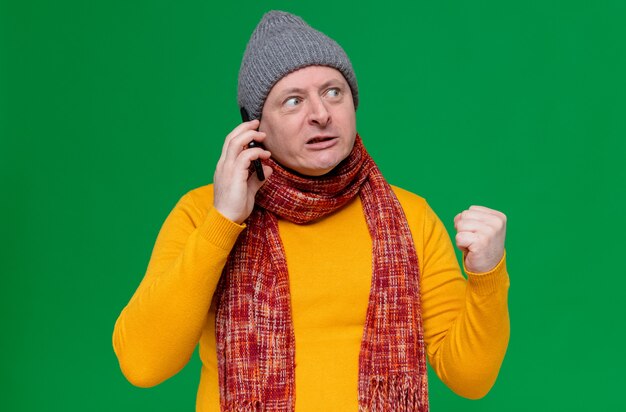 Hombre eslavo adulto molesto con gorro de invierno y bufanda alrededor de su cuello hablando por teléfono y manteniendo el puño mirando a un lado