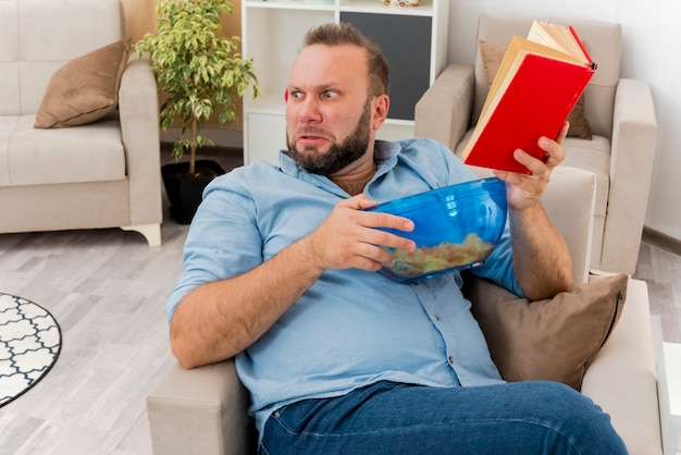 Hombre eslavo adulto molesto se sienta en un sillón sosteniendo un tazón de patatas fritas y un libro mirando de lado dentro de la sala de estar