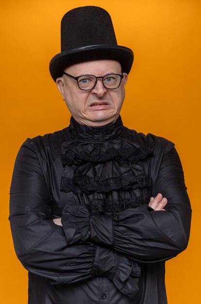 Hombre eslavo adulto molesto con sombrero de copa y gafas ópticas en camisa gótica negra de pie con los brazos cruzados