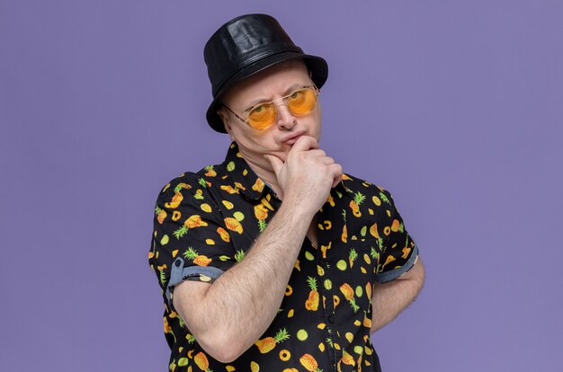 Hombre eslavo adulto reflexivo con sombrero de copa negro con gafas de sol poniendo la mano en la barbilla y