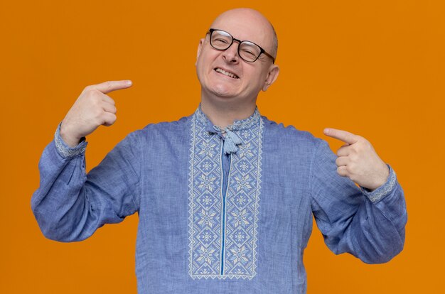Hombre eslavo adulto sonriente en camisa azul y con gafas ópticas apuntando a sí mismo