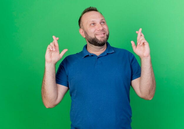 Hombre eslavo adulto sonriente mirando al lado cruzando los dedos aislado en la pared verde