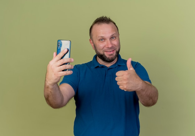 Hombre eslavo adulto sonriente sosteniendo teléfono móvil y mostrando el pulgar hacia arriba