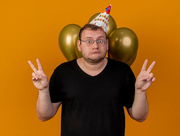 Hombre eslavo adulto sorprendido con gafas ópticas con gorro de cumpleaños se encuentra frente a globos de helio gesticulando el signo de la mano de la victoria con las dos manos