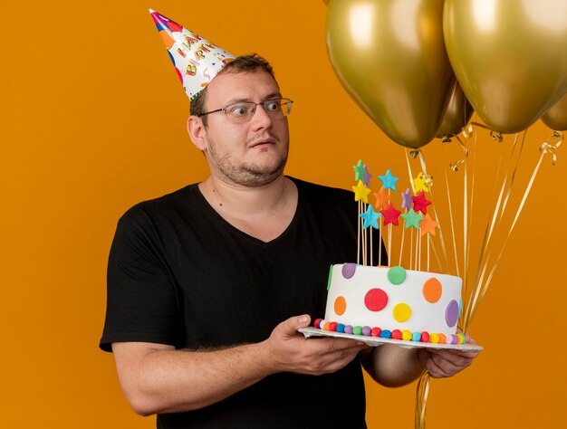 Hombre eslavo adulto sorprendido con gafas ópticas con gorro de cumpleaños sostiene globos de helio y pastel de cumpleaños