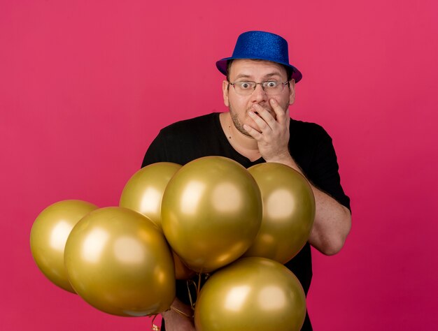 Hombre eslavo adulto sorprendido con gafas ópticas con sombrero de fiesta azul pone la mano en la boca y sostiene globos de helio