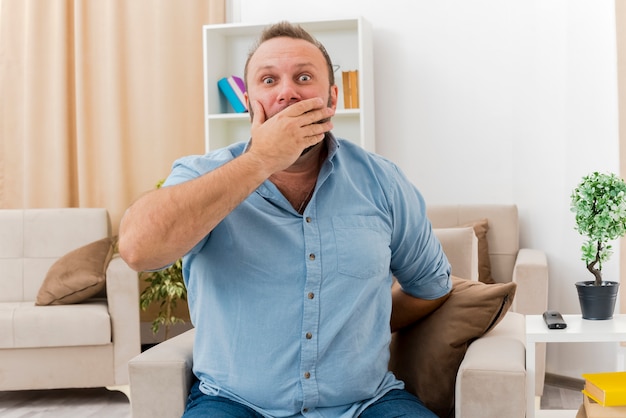 Hombre eslavo adulto sorprendido se sienta en el sillón poniendo la mano en la boca y sosteniendo la mano desde atrás dentro de la sala de estar