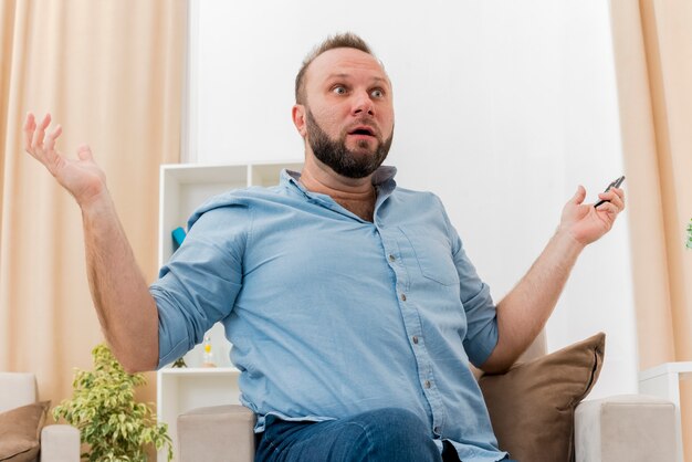 Hombre eslavo adulto sorprendido se sienta en un sillón sosteniendo el control remoto de la televisión con las manos abiertas dentro de la sala de estar