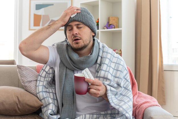 Hombre eslavo enfermo dolorido con bufanda alrededor del cuello con sombrero de invierno poniendo su mano en la cabeza y sosteniendo una taza con blister de medicamentos sentado en el sofá en la sala de estar