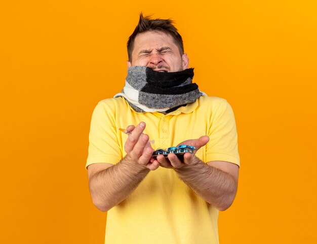 Hombre eslavo enfermo rubio joven disgustado que cubre la boca con bufanda sostiene paquetes de píldoras médicas y jeringas aisladas en la pared naranja con espacio de copia