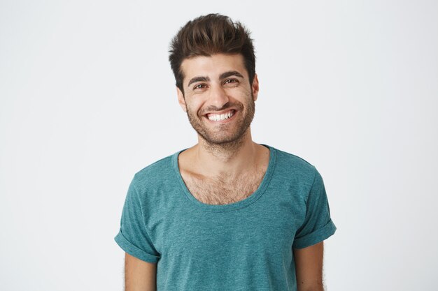 Hombre español positivo y alegre con barba con camiseta azul sonriendo alegremente sintiéndose feliz por el día libre en el trabajo. Concepto de personas y emociones