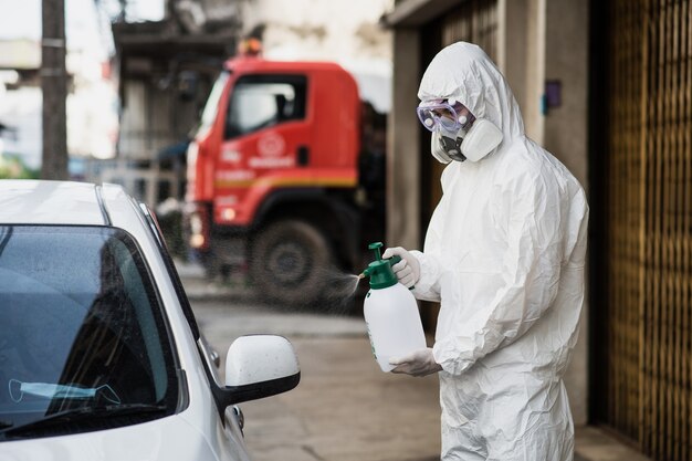 Hombre especialista en desinfección con traje de equipo de protección personal (EPI), guantes, máscara y gafas transparentes, limpiando el automóvil con una botella de desinfectante en aerosol presurizado para eliminar el covid-19