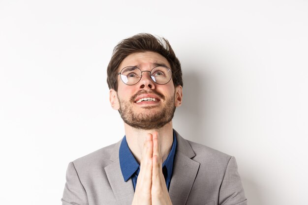 Hombre esperanzado nervioso con gafas y traje rogando a Dios, pidiendo por favor y estrechándole la mano en oración, fondo blanco.