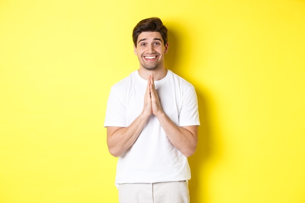 Hombre esperanzado pidiendo ayuda, pidiendo favor, necesita algo y sonriendo, de pie sobre un fondo amarillo.