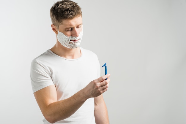 Hombre con espuma de afeitar en su cara mirando la maquinilla de afeitar azul de pie contra el fondo gris