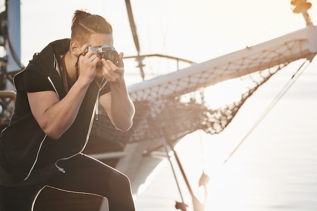 El hombre está buscando las mejores fotos. Fotógrafo enfocado durante el trabajo parado cerca de la flexión del yate mientras mira a través de la cámara, toma fotos del mar o del puerto, toma fotos con el concepto de estilo de vida