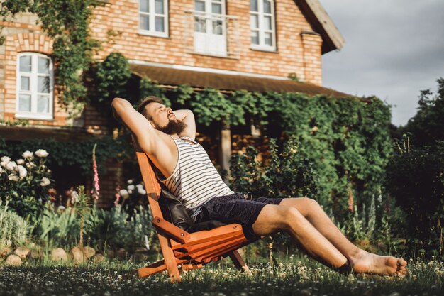 un hombre está descansando en una casa de campo. Un hombre barbudo disfruta de la puesta del sol en un césped verde.