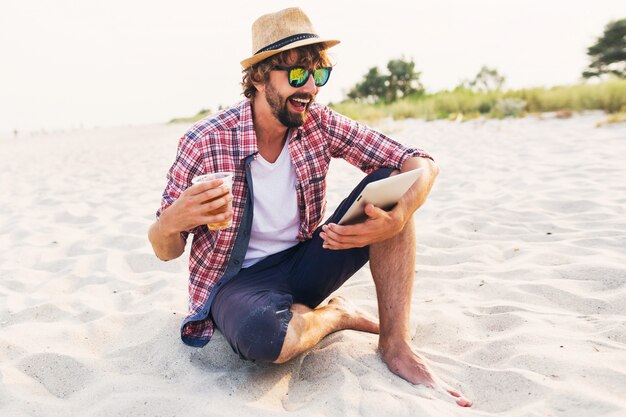 Hombre con estilo feliz usando tableta y bebiendo cerveza en la playa