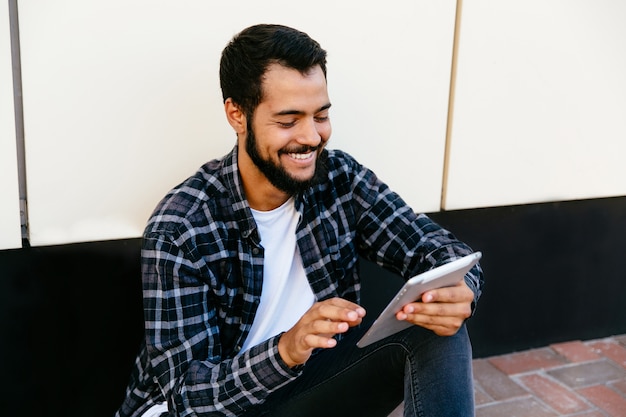 Hombre con estilo hipster joven usando una tableta digital, sonriendo alegremente, mirando la pantalla de la tableta