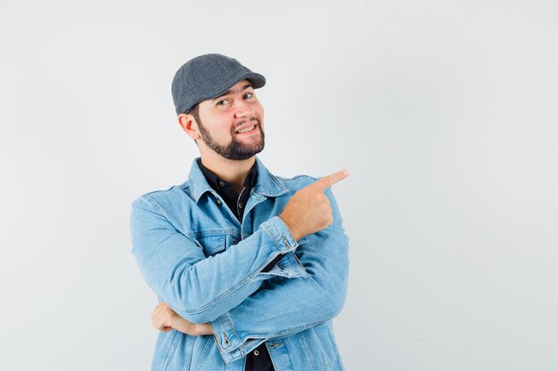 Hombre de estilo retro apuntando a la izquierda con chaqueta, gorra, camisa y mirando feliz, vista frontal.