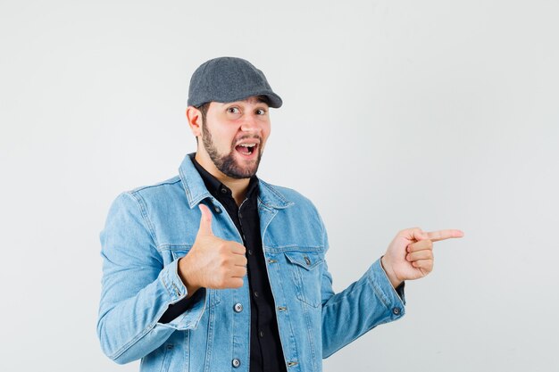 Hombre de estilo retro apuntando a la izquierda mientras muestra el pulgar hacia arriba en chaqueta, gorra, camisa y se ve alegre. vista frontal.