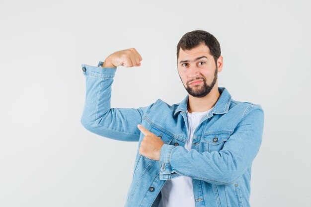 Hombre de estilo retro apuntando a los músculos de su brazo en chaqueta, camiseta y mirando satisfecho de sí mismo, vista frontal.