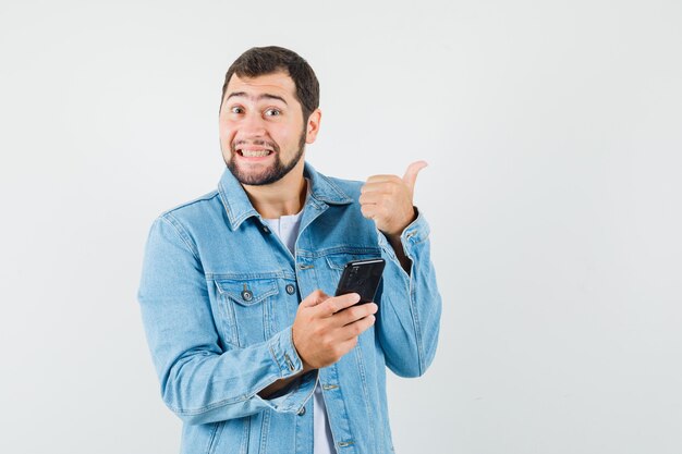 Hombre de estilo retro con chaqueta, camiseta apuntando hacia afuera mientras sostiene el teléfono y se ve alegre, vista frontal.