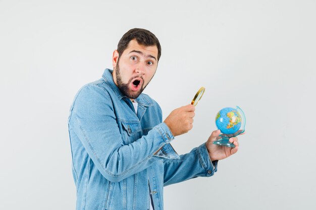 Hombre de estilo retro con chaqueta, camiseta mirando el mini globo con lupa y mirando sorprendido.