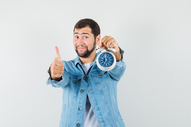 Hombre de estilo retro con chaqueta, camiseta mostrando el pulgar hacia arriba mientras sostiene el reloj y se ve satisfecho, vista frontal.