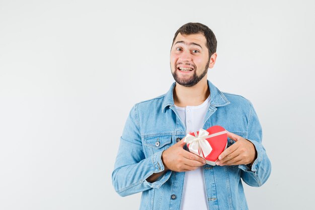 Hombre de estilo retro con chaqueta, camiseta sosteniendo una mini caja de regalo y mirando alegre, vista frontal. espacio para texto