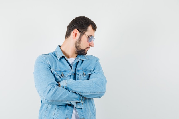 Hombre de estilo retro con chaqueta, gafas de sol mirando a un lado mientras está de pie con los brazos cruzados, vista frontal.