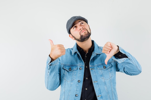 Hombre de estilo retro con chaqueta, gorra, camisa mostrando el pulgar hacia arriba y hacia abajo y mirando confundido, vista frontal.