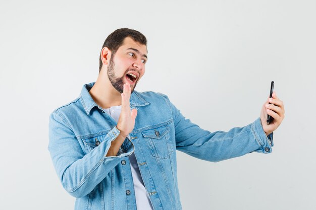 Hombre de estilo retro que muestra un gesto de saludo mientras hace una videollamada en chaqueta, camiseta y parece concentrado. .