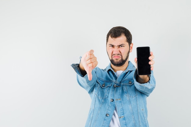 Hombre de estilo retro que muestra el pulgar hacia abajo mientras muestra el teléfono móvil con chaqueta, camiseta y aspecto negativo. vista frontal.