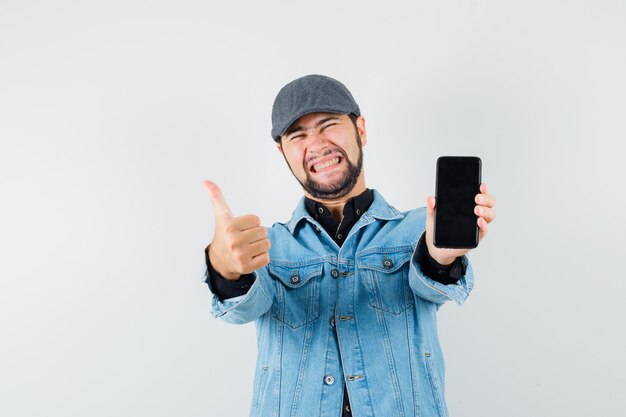 Hombre de estilo retro que muestra el pulgar hacia arriba mientras muestra el teléfono en la chaqueta, gorra, camisa y se ve feliz, vista frontal.