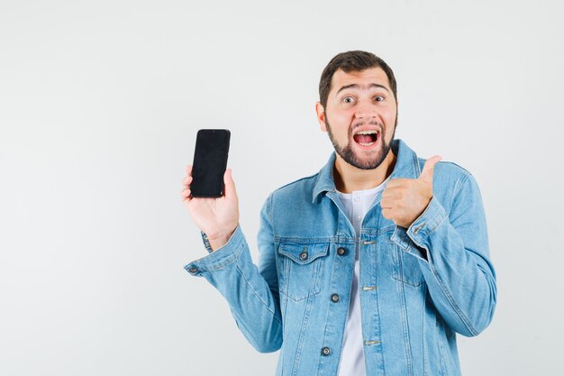 Hombre de estilo retro que muestra el pulgar hacia arriba mientras sostiene el teléfono en la chaqueta, camiseta y se ve complacido, vista frontal.