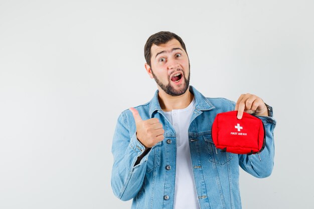 Hombre de estilo retro que sostiene el botiquín de primeros auxilios mientras muestra el pulgar hacia arriba en la chaqueta, camiseta y parece optimista. vista frontal.