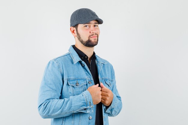 Hombre de estilo retro sosteniendo su cuello en la chaqueta, gorra y mirando confiado, vista frontal.